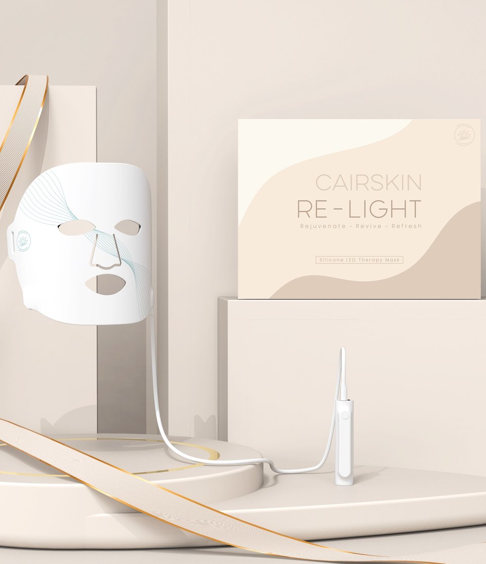 White CAIRSKIN RE-Light silicone LED mask on an ivory pedestal with its box and a gold-edged ribbon