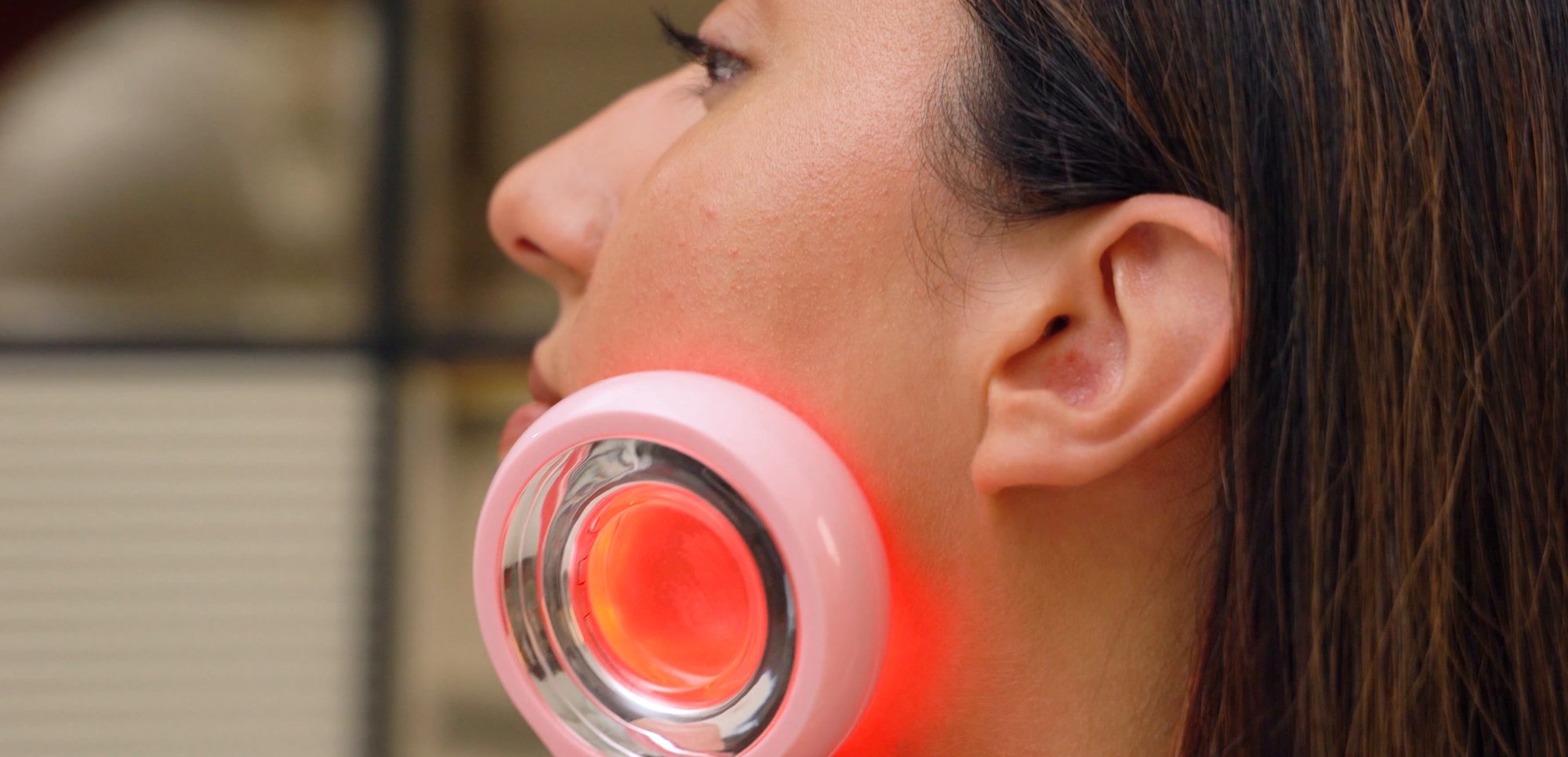 Side profile of a woman holding the pink CAIRSKIN Collagen Booster against her jaw, its ring of red light glowing
